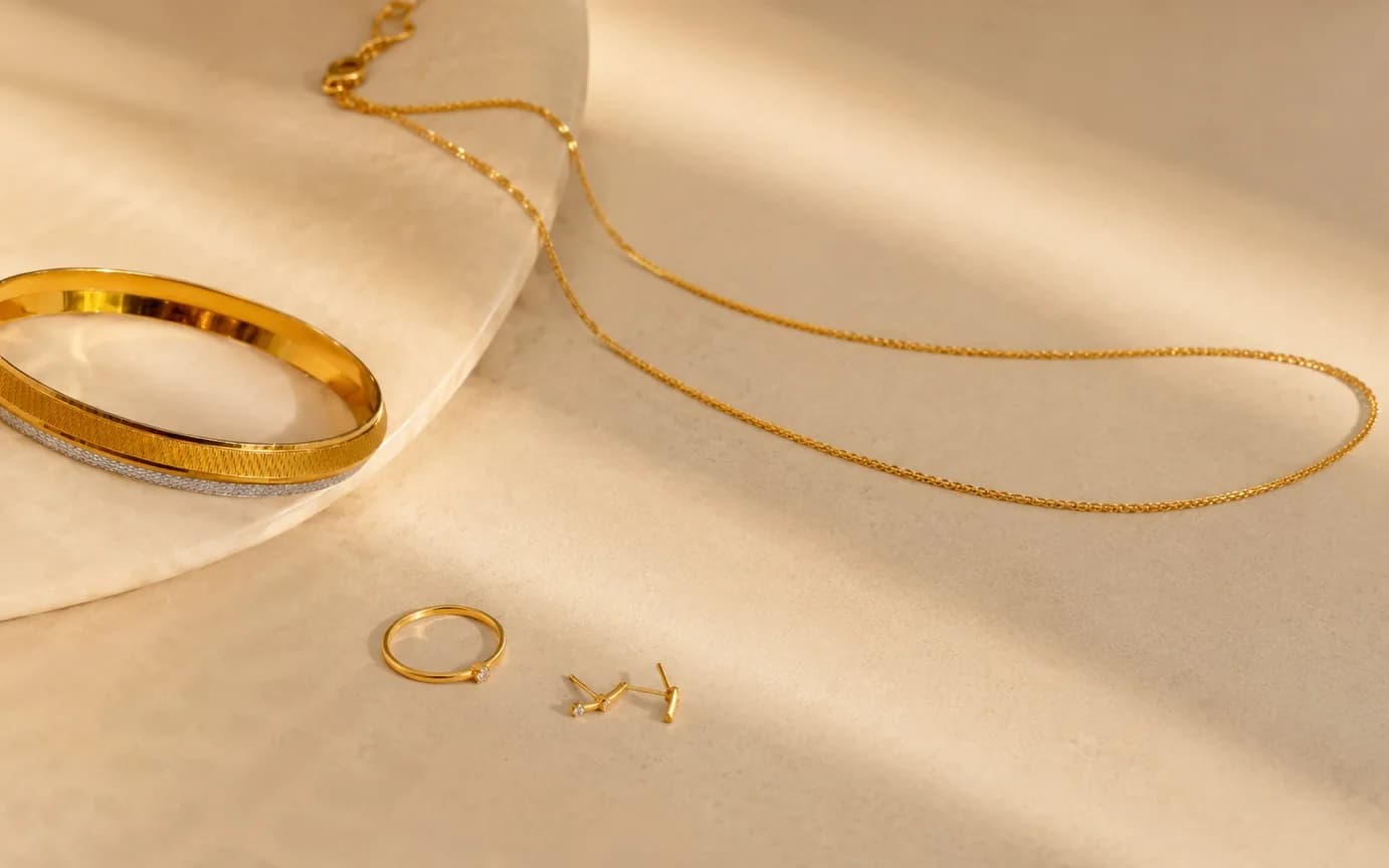 A slim gold bangle, a fine chain and a plain band laid out on a pale stone slab.